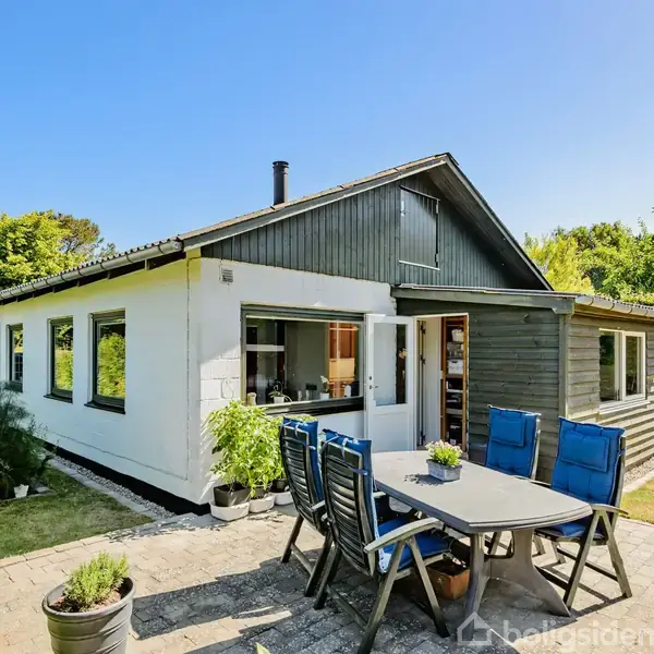 Hus med hvid facade og mørkt trætag står i en grøn have. Terrasse med bord og blå stole foran huset. Solrig dag med klar blå himmel.