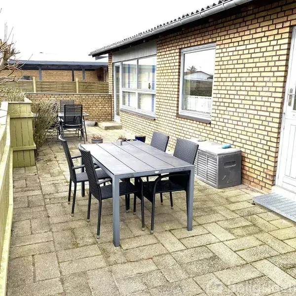 Terrassemøbler står på flisebelagt terrasse ved et murstenshus. Seks stole omringer et bord, mens en hæk omkranser terrassen på venstre side. En havedør fører ind i huset.