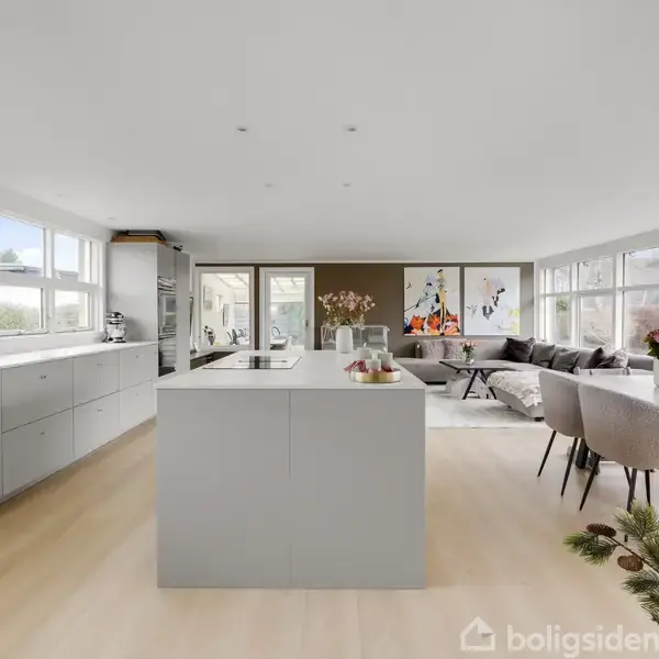 Et moderne køkkenalrum med en stor ø i midten, omgivet af grå skabe. I baggrunden findes en spiseplads med et bord samt stole og kunst på væggen. Lyse trægulve og store vinduer.