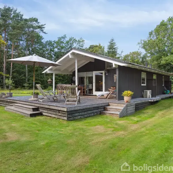 Et sort sommerhus med hvide detaljer står på en grøn græsplæne. Et træterrasseområde foran huset har havemøbler og en stor parasol. Omgivet af høje træer.