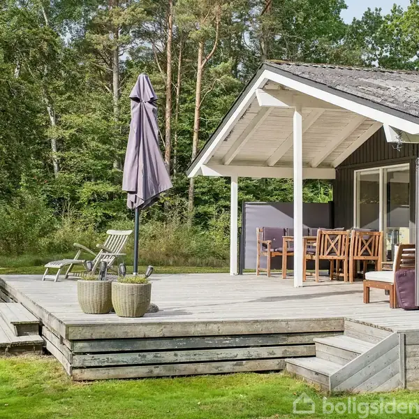 Sort træhus med overdækket terrasse, hvor der står træmøbler og parasol. Terrassen er hævet, har trapper og står i en grøn have omgivet af skovbevoksning.