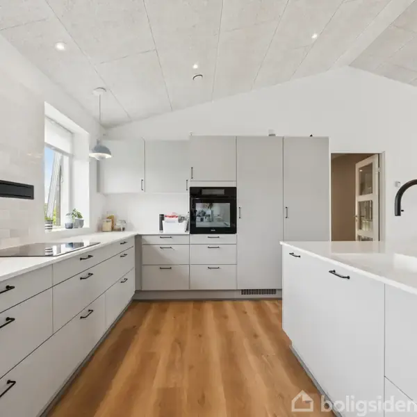 Et moderne køkken med hvide skabe og skuffer, sort håndvask og trælaminatgulv. Køkkenet indeholder indbyggede apparater og er omgivet af lyse vægge og et vindue med udsigt.