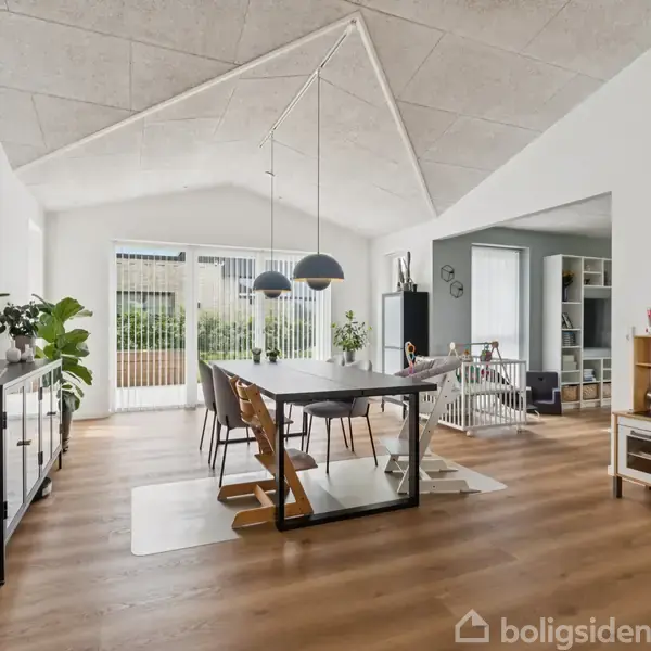 Et spisebord står centralt i et lyst rum med store vinduer, omgivet af planter og hylder. En babygård og en legekøkken er placeret i baggrunden på trægulvet.