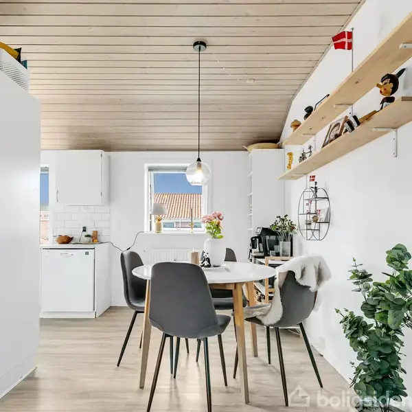 Rundt spisebord med grå stole står i en lys spisestue med trægulv og træloft. Der er planter, hylder med dekorationer og et stort vindue i baggrunden, der giver naturligt lys.