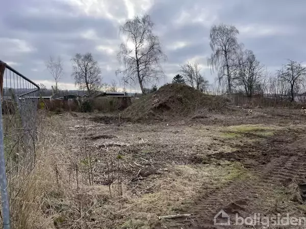 En jordhøj ligger i en åben, tilgroet mark, omgivet af rustne træer under en overskyet himmel. En metalhegn ses til venstre. Vandmærke "Boligsiden" ignoreres.