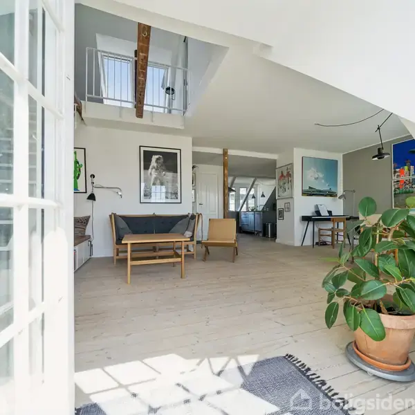 Stue med sofa og lænestol står stille på trægulv. Store vinduer lader sollys strømme ind. Grøn plante i forgrunden og kunstværker på væggene skaber en lys atmosfære.