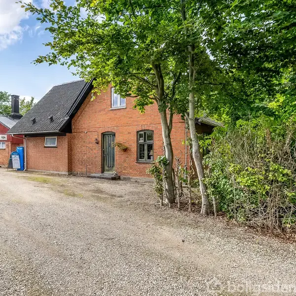 Een rødstenshus står på en grusvej, omgivet af frodige træer og buske. I baggrunden ses en mindre, rød bygning. En blå beholder er placeret tæt på huset.