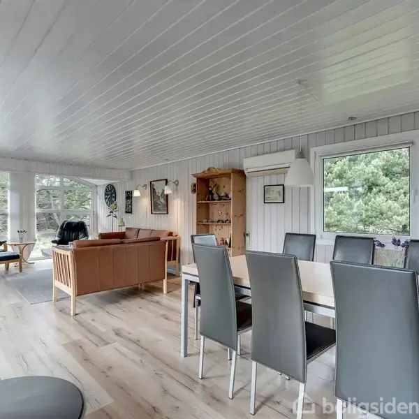 En moderne stue med brændeovn brænder ild, og et spisebord med høje, sorte stole i en lys træbeklædt stue med store vinduer og udsigt til grønne træer.
