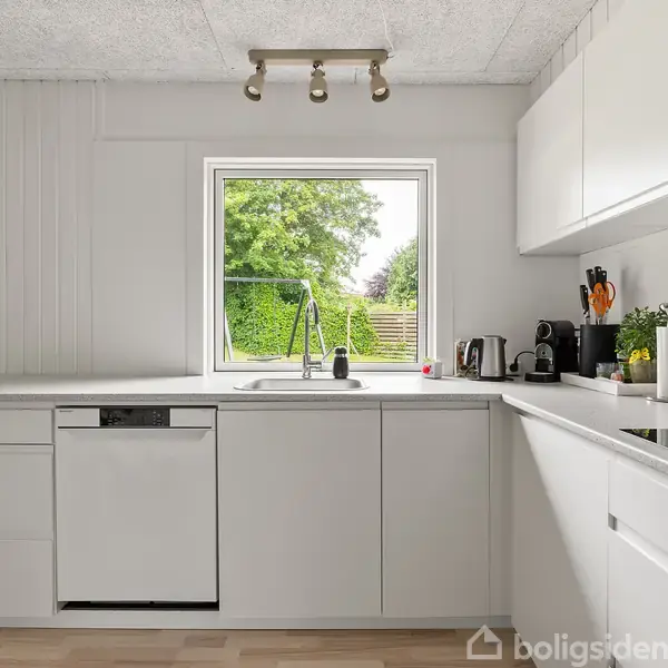 Et hvidt, moderne køkken med indbygningsskabe og hvidevarer. Et vindue over vasken viser en grøn have. Bordpladen har kaffemaskine, planter og opbevaringskrus.