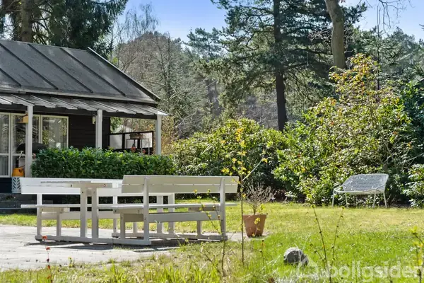 Træbænk med bord står på en flisebelagt terrasse foran et hus med grønne planter og buske i baggrunden, omgivet af en frodig have og høje træer.
