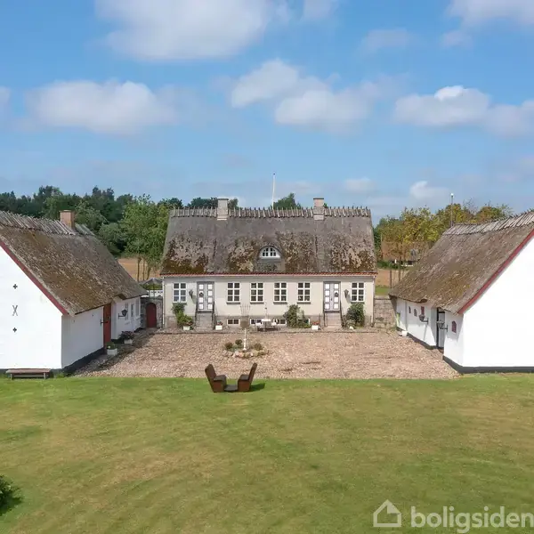 Et bindingsværkshus med stråtækt tag står mellem to mindre bygninger. Handling: Det er omgivet af en gårdsplads. Kontekst: Grønne græsarealer og træer i baggrunden under en blå himmel.
