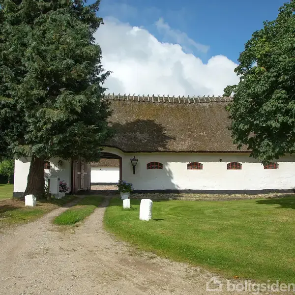 Et hvidt stråtækt hus står omgivet af grønne træer og en grusvej i en landlig have. Skyer og klar himmel ses i baggrunden.