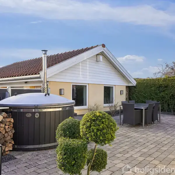 En udendørs terrasse med et rundt vildmarksbad i forgrunden, omgivet af stablet brænde. En gul bygning med rødt tag ses i baggrunden, samt et spisebord med stole.