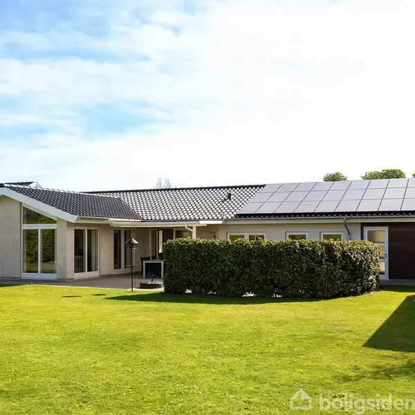 Et moderne hus med solpaneler på taget står omkranset af en velplejet græsplæne. Lave hække skaber afskærmning ved indgangen. En klar blå himmel ses i baggrunden.