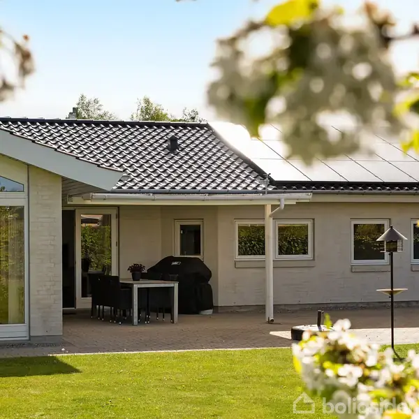 Et moderne hus med solpaneler på taget står med en terrasse og havemøbler. Træer med blomster omkranser græsplænen, hvilket skaber en idyllisk haveatmosfære.