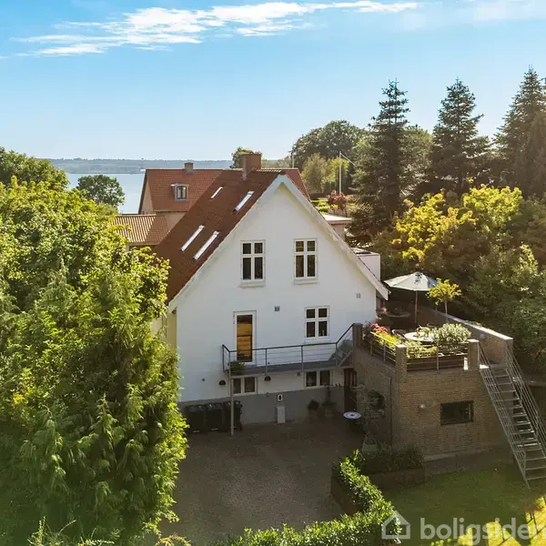 Hus med hvid facade og rødt tag står i centrum, omgivet af træer og en terrasse. I baggrunden ses en sø eller hav.