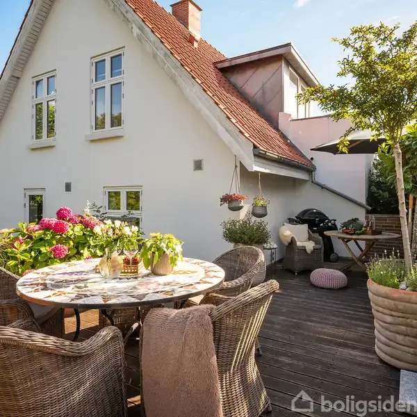 Et rundt bord med kurvestole omgivet af farverige blomster på en terrasse foran et hvidt hus med skråt tag. Solskin og grønne planter skaber en indbydende atmosfære.