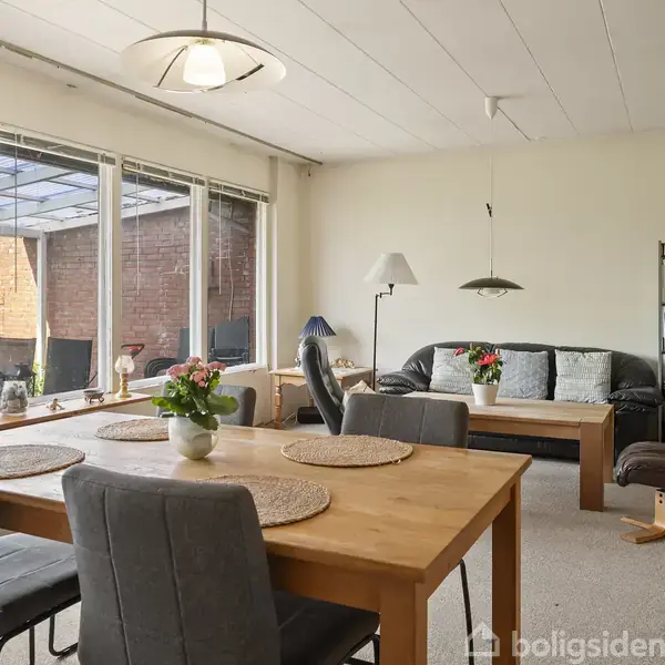 Spisebord med fire stole står i stuen, der modtager lys fra store vinduer med udsigt til terrasse. Sofa og reol er i baggrunden, hvilket skaber en hyggelig atmosfære.