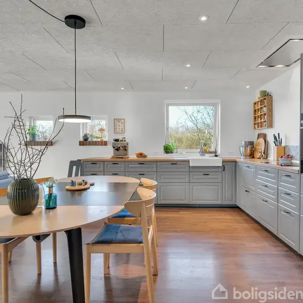 Et moderne køkken indeholder grå skabe og indbygget ovn; et ovalt spisebord med stole står centralt, mens en række planter og dekorationer skaber hygge. Et vindue viser udsigt udenfor.