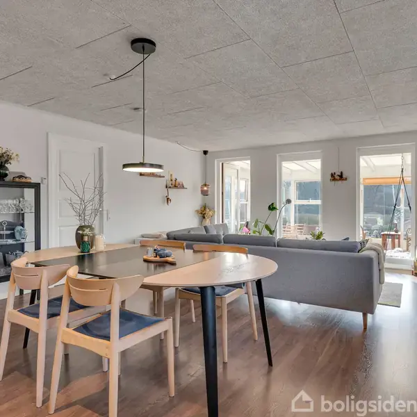 Spisebord med fire stole er centralt placeret i en stue. Baggrunden viser en grå sofa ved store vinduer, lysindfald og en guitar hængende på væggen.