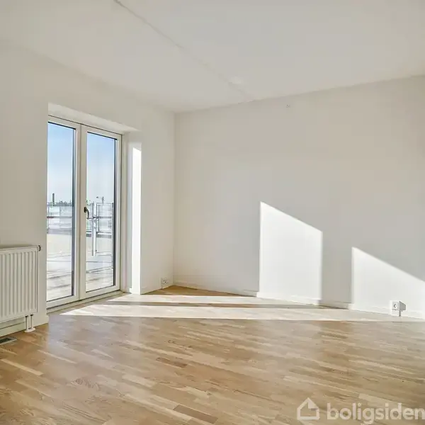 Et lyst værelse med trægulv og hvide vægge. Sollys strømmer ind gennem dobbeltdøre, der fører til en balkon. Der er en radiator ved siden af dørene.