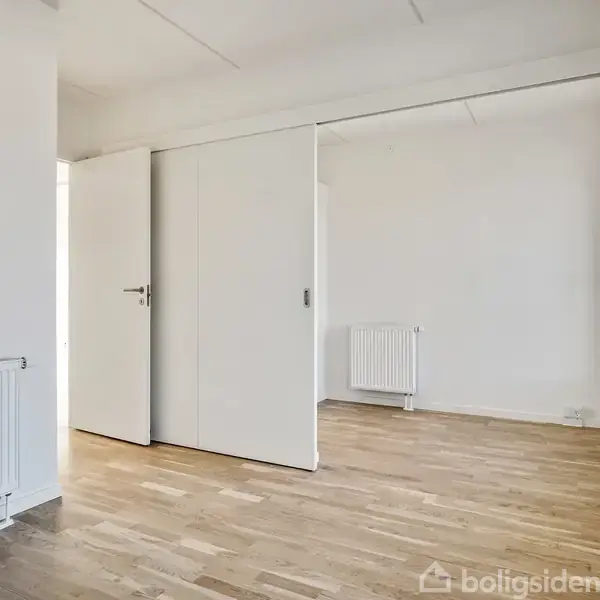 Et tomt værelse med trægulv har åbne skydedøre, der fører til et andet rum. To radiators er synlige, en i hvert rum, med lyse vægge og et lyst loft.