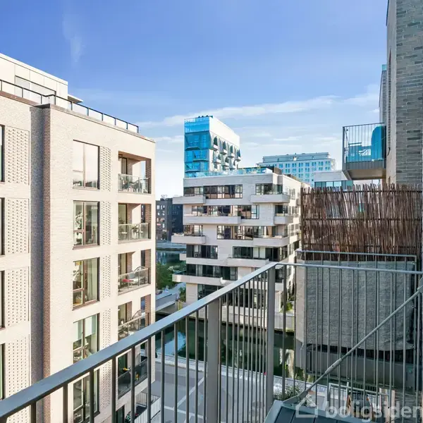En altan med metalrækværk strækker sig ud fra en lejlighed i en storby. Omgivet af moderne lejlighedsbygninger under en klar blå himmel.