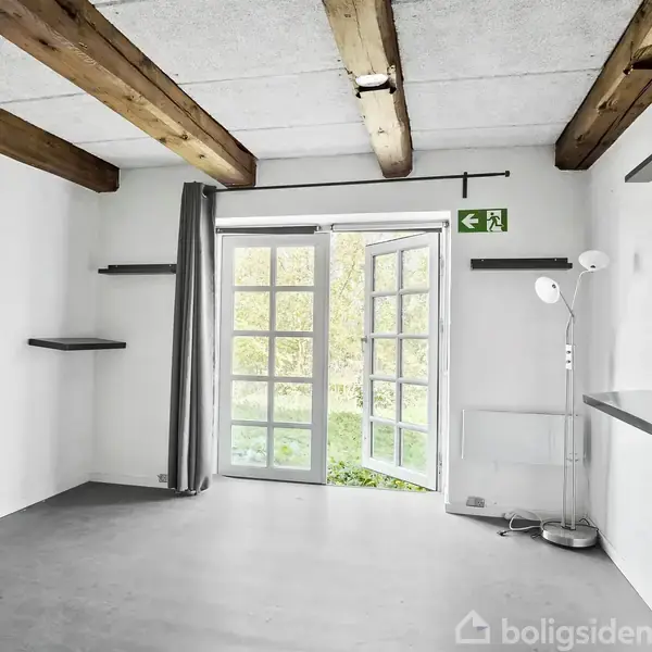 En tom stue med trælofte, har åbne dobbeltdøre ud til en have, grøn nødudgangsskilt ovenfor. Rummet har hylder på væggene og en stående lampe i hjørnet.