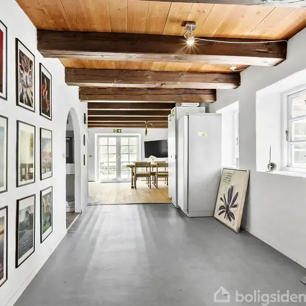 En gang med trægulv og synlige loftsbjælker er dekoreret med ni kunstværker på venstre væg. I baggrunden ses et lyst rum med store vinduer og et spisebord.