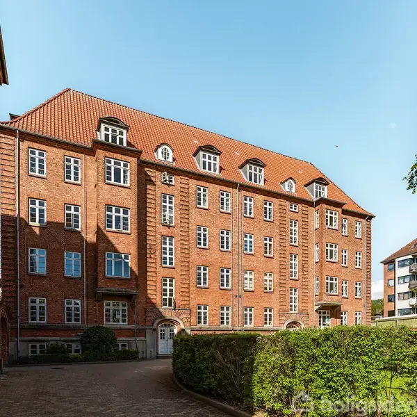 Rødt murstensbygning med mange vinduer står stille. Bygningen har et rødt tag med kvistvinduer. Den er omgivet af grønne hække og ligger i et solrigt område med en klar blå himmel.