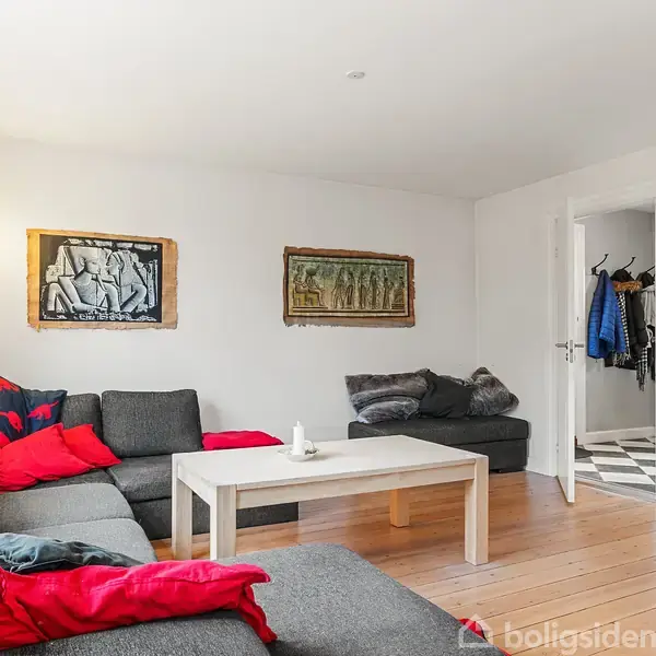 En grå hjørnesofa med røde puder står i en stue med lyse trægulve. Et hvidt sofabord står foran sofaen, og væggen har to kunstværker. En fjernsynsskærm hænger til højre.