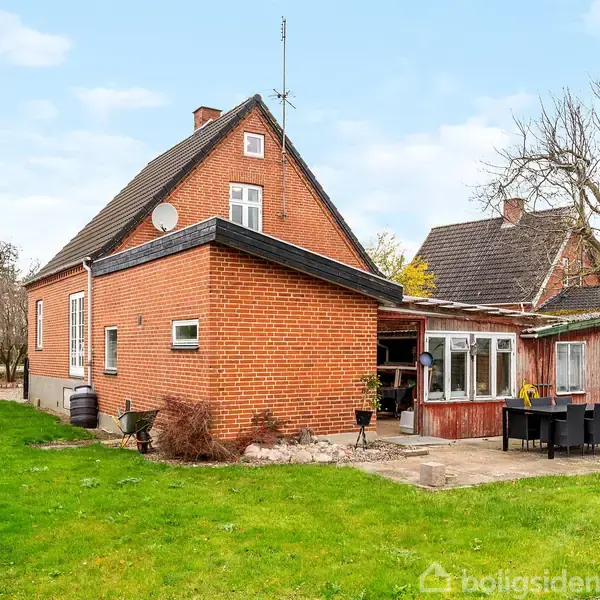 Et rødt murstenshus med en trekantet gavl står stille i en have, omgivet af grønne plæner og træer. Ved siden af huset er der en terrasse med et spisebord.