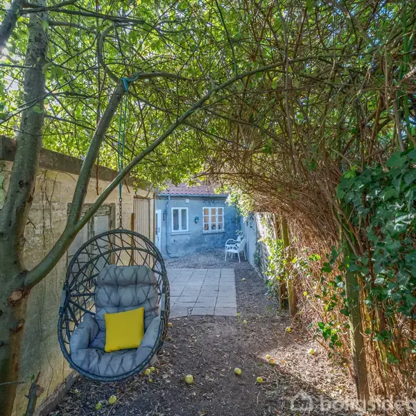 Spisebord med hvide stole står i lyst rum med trægulv. En orange lampe hænger over bordet, og der er et farverigt maleri på væggen. En hylde, sofa og et lille bord ses i baggrunden. Åben forbindelse til køkkenet.