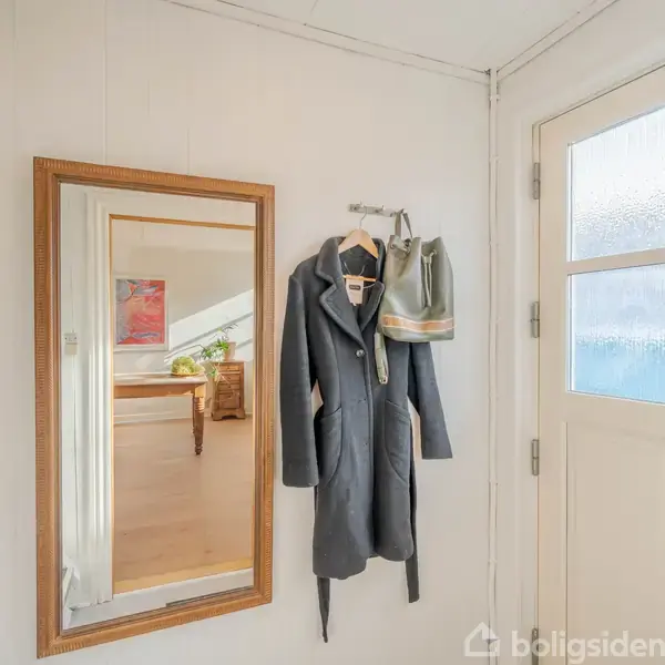 En frakke hænger på en knage ved siden af en grå taske. En stor træindrammet spejl hænger på væggen i en lys entré ved siden af en dør med matterede glasruder.