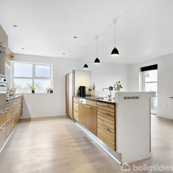 Et moderne køkken med træskabsfronter og hvidevarer, herunder en indbygget ovn. Lyse pendellamper hænger over køkkenøen. Rummet har store vinduer, der giver naturligt lys.
