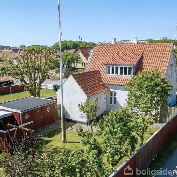 Et hvidt hus med rødt tegltag står blandt grønne træer og buske i en villakvarter. En rød trægarage er placeret ved siden af huset. En gammel vindmølle ses i baggrunden.