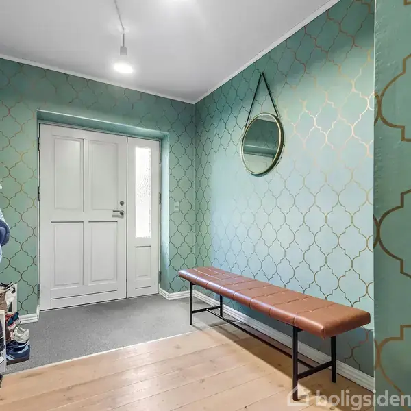 Bænk med brun sæde står stille i en entré med grøn, mønstret tapet. Rundt spejl hænger på væggen over bænken. Til venstre opbevares overtøj og sko.