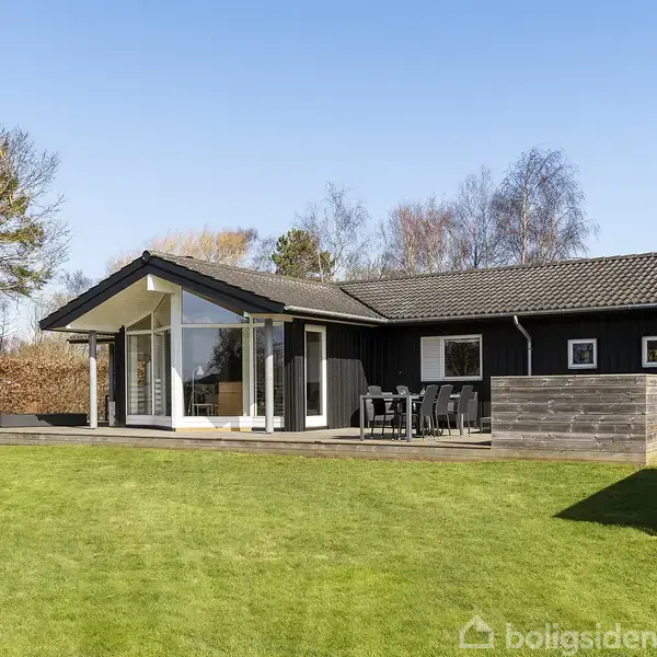 Et sort enfamiliehus står på en velplejet græsplæne. Der er en terrasse med havemøbler foran huset. Omkring er der træer og en blå himmel.