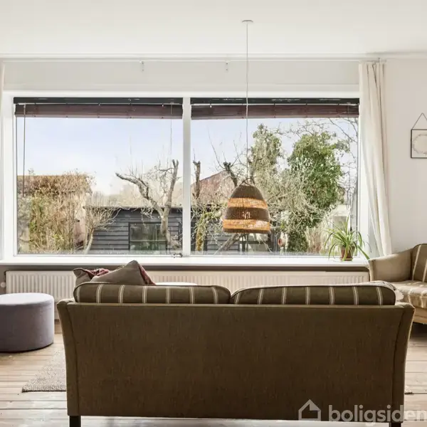 En stue med en sofa og to stole vendt mod et stort vindue. Udenfor ses en have med træer. Bøgerne på reolen og en lampe danner en hyggelig atmosfære.
