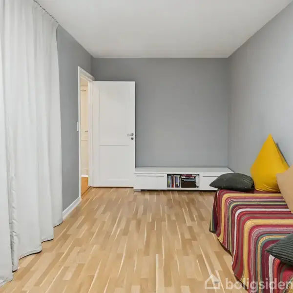 Sofa med stribede puder står langs en væg i et rum med grå vægge og lyst trægulv. En hvid dør og bogreol ses i baggrunden.