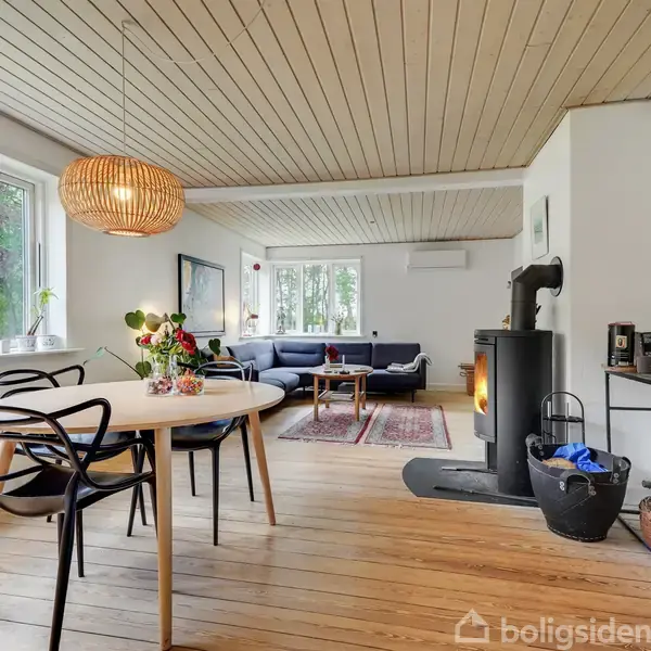 En stue med spisebord og sorte stole, en brændeovn i hjørnet, sofaområde i baggrunden. Store vinduer giver naturligt lys, og der er planter og dekorationer på bordet.