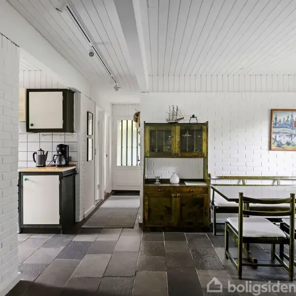 Et spisestue med et bord og seks stole står i et rum med hvidmalede murstensvægge. Et vitrineskab rummer porcelæn, og et moderne lampe hænger fra loftet.