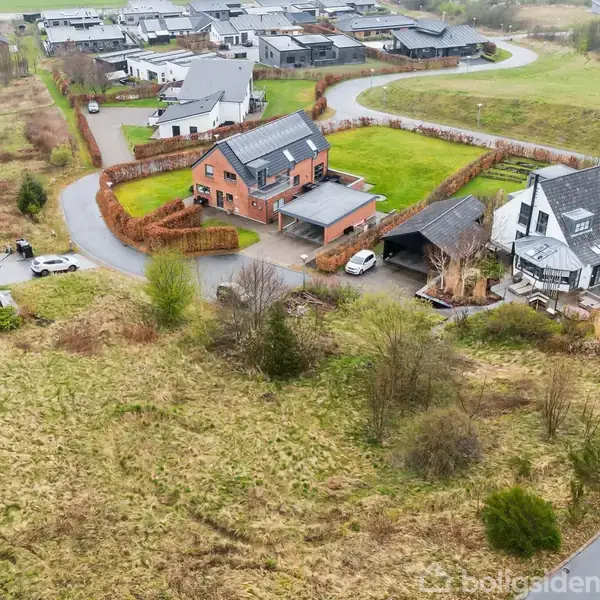 Flere huse står i et grønt, kuperet landskab. Et murstenshus med grå tag ligger ved siden af et sort-hvidt hus med bil i indkørslen. Græsarealer omkranser området.