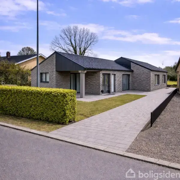En moderne bungalow i grå mursten med fladt tag står på en velholdt grund. Foran huset er en flisebelagt indkørsel, omgivet af grønne hække og en lygtepæl i kanten.