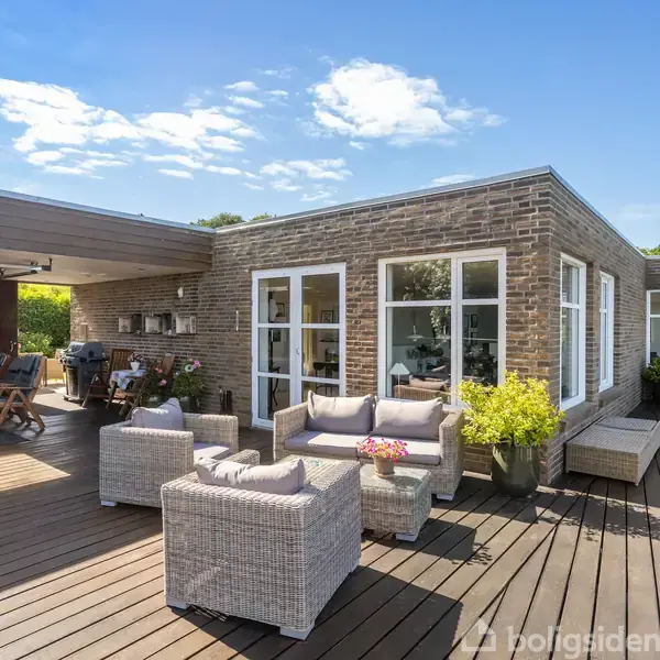 En moderne terrasse med polyrattan møbler står foran et hus med store glasvinduer. Terrassebordet og stole under en halvtag til venstre skaber en hyggelig udendørs spiseplads.