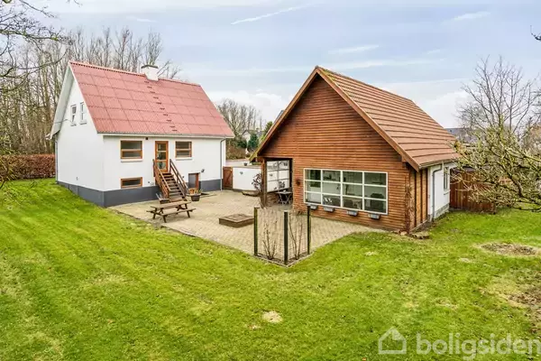 To huse står i en have. Det ene er hvidt med rødt tag, det andet i træ med store vinduer. Mellem dem er en terrasse med et picnicbord.