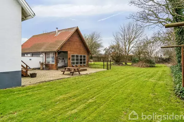 Et træhus med rødt tag står på en velplejet græsplæne. Et picnicbord er placeret på en flisebelagt terrasse foran huset. Omkringliggende træer markerer grunden.