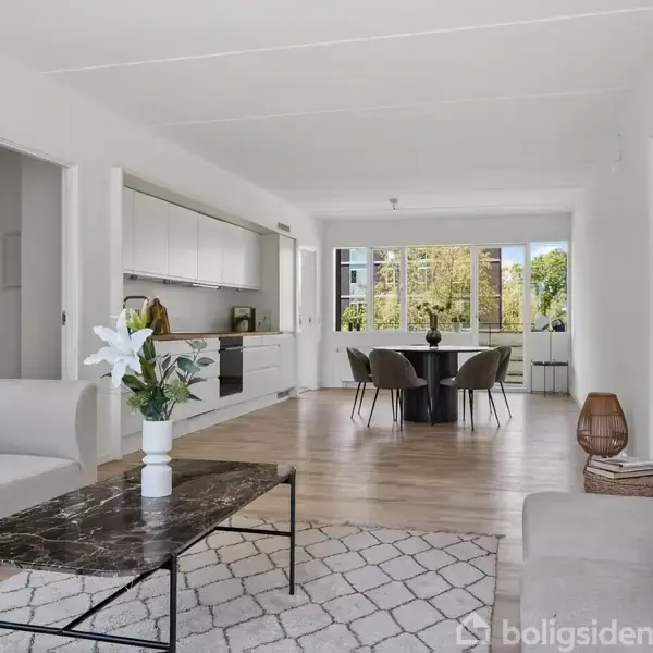 En lys, moderne stue med en sort marmorbord, sofaer på hver side, og en hvid vase med blomster. Køkkenet ligger i baggrunden, med en spisestue foran store vinduer.