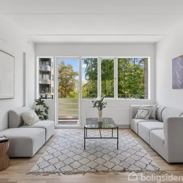 Stue med to grå sofaer omkring et lille sofabord. En stor vinduesfront viser grønne træer udenfor. Rummet er lyst og moderne, med et tæppe og minimalistisk indretning.