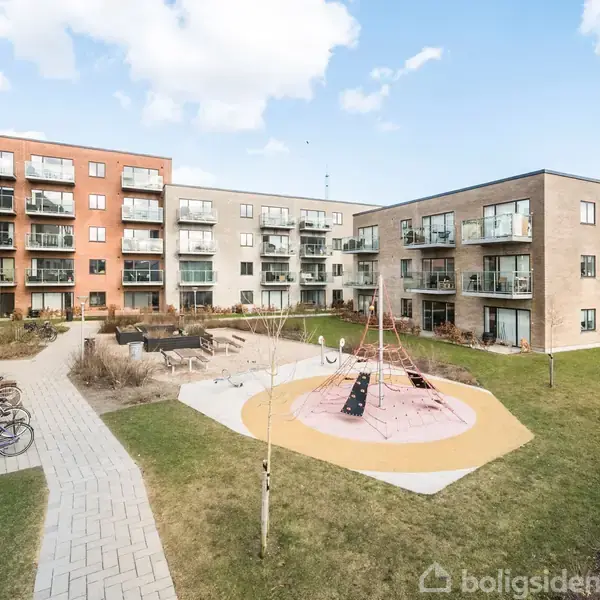 Legeplads med klatrestativ står midt i en gårdhave omgivet af moderne lejlighedsbygninger. Flisebelagte stier løber langs grønne områder, og cykler er parkeret langs stien.